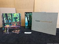 Two photographic books displayed side by side showing their covers: "At Home Canada" with a colorful nature and village scene on cover and "Canada A Year of the Land" with grey cover and gold lettering.
