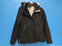 Front view of black ladies' ONYX ski jacket with zipper and pockets, hanging against a blue background.