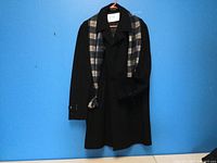Full view of men’s black dress coat hanging on hanger, with plaid scarf draped over it.