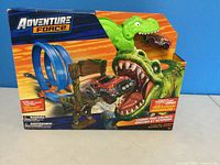 Front view of the toy packaging showing the blue double loop track, green dinosaur figure, and red-black vehicle inside.