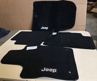 Full set of four black carpeted Jeep floor mats displayed on a table, showing two front mats and two rear mats with Jeep logo and securing holes.