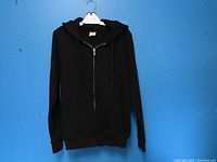 Full view of black men's zipper hoodie hanging on a hanger against a blue background.