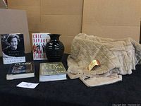 Image showing a beige microfibre throw, black ceramic vase, and five hardcover books arranged on a black surface with cardboard background.