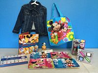 Jean jacket size 4 dark blue on a hanger next to Mickey Mouse themed tote bag, puzzle, placemats, small toy figure, book and markers set on table against blue wall.