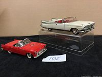Two vintage car model cars on display stand, one white convertible with red interior and one red convertible with chrome details.