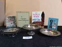 Four cookbooks and three silver-tone serving dishes displayed together on a table