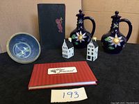View of full lot including two blue floral ceramic decanters, vintage Esquire's Handbook for Hosts, red wine journal, two white ceramic collectible houses, and a blue glazed candy dish bowl on black surface.