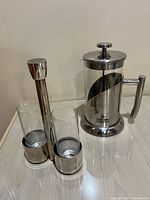 Photo showing Crio Bru French press beside two glass cups in stainless steel holder on a reflective surface.
