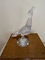 Front and side angles of transparent and frosted glass bird statue standing on round base. Beak is visibly chipped at close range.