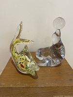 Two glass fish sculptures placed side by side on wooden surface, one clear and one colorful Murano style.
