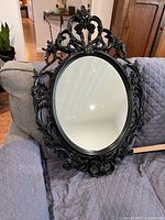 Full view of the ornate oval mirror resting on a couch, showing the detailed baroque style black plastic frame and clear mirror surface.