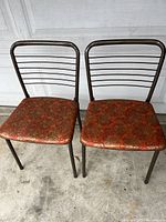Pair of mid-century modern folding chairs with brown taupe metal frames and vintage vinyl patterned seats shown front view.