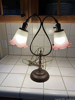 Vintage two-light table lamp on tiled counter, lit with frosted glass tulip-shaped shades with pink edges, brass base with patina.