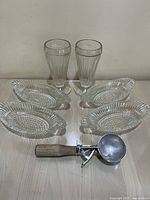 Photo of 4 clear glass banana split dishes, 2 clear tall milkshake glasses, and 1 aluminum scoop with wooden handle on a table.