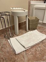 White plastic stool, metal toilet paper roll holder, beige textured waste basket, two beige towels arranged on tiled floor