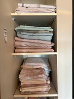 Three shelves containing neatly folded bed linens, sheets, and pillowcases in various pastel tones. Top shelf has light colored linens; middle shelf has blue and pink sheets; bottom shelf has pink and floral patterns.