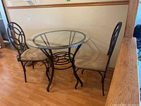Photo of bistro table with round glass top and two matching metal chairs with beige cushions against wall on wood floor.