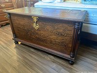 Side view of dark wood carved blanket chest showing scenic rustic relief carvings and turned corner columns.