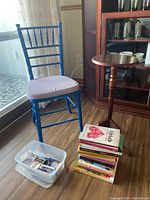 Blue Chiavari chair next to wooden pedestal stand, with books stacked and a plastic bin of cassette tapes on the floor