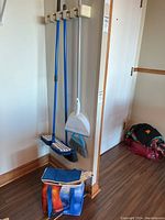 Photo showing two blue-handled brooms, one blue-handled mop, white dustpan with brush hanging on a wall rack, and a blue bag on the floor containing light bulbs.