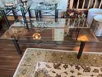 Full view of the rectangular glass top coffee table with black metal base on a patterned rug, showing the clear glass and structure underneath.