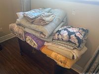 Stack of various bedding items on wooden furniture including comforters, blankets and mattress topper with floral and solid colors visible.