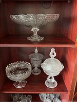 Photo of four crystal dishes on two wooden shelves; top shelf holds large fruit bowl, bottom shelf holds two candy jars with lids and one footed compote dish.