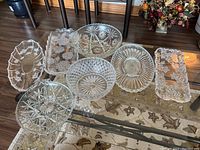 Wide shot of nine assorted crystal and glass platters, serving dishes and bowls placed on a carpeted floor under a glass table.
