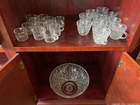 Shelved display showing 22 glasses total, 10 matching crystal glasses with a detailed cut starburst pattern and 12 plain clear glass cups, all arranged on a dark wood shelf. The crystal punch bowl is visible below the shelf inside a cabinet.