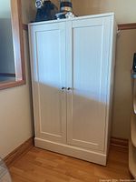Full front view of the white painted wood cupboard standing on a wooden floor next to a wall