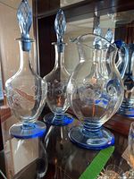 Photo showing two clear etched glass decanters with blue bases and stoppers alongside a matching etched glass pitcher with a blue base.