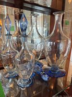 Four etched glass decanters with blue stoppers and bases, and one matching pitcher with etched design and blue base, shown inside a wooden display cabinet.