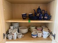 Full display inside a wooden cupboard showing two shelves with various cups, mugs, and saucers including the blue ceramic tea set.