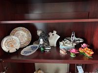 Shelf showing decorative plates, Buddha and abstract figurines, ceramic flowers and small animal figurines
