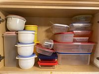 View of multiple plastic and glass food storage containers stacked in cabinet, showing various sizes and colored lids.