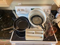 Photo showing frying pans stacked on stove surface, plastic colander filled with mixing bowls and glass measuring cups arranged beside them, with white plastic cutlery organizer in front.