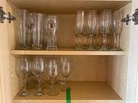 Tall beer glasses including one with Alexander Keith's logo, clear champagne flutes, and clear wine glasses arranged on shelves in a wooden cabinet.