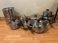 Front view of seven stainless steel cookware pots with glass lids and black handles arranged on wooden floor.