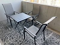 Outdoor patio set on balcony, includes one black square metal frame table with slatted top and three matching patio chairs with light gray plastic mesh seating and metal armrests.