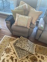 Five throw cushions including a beige textured, a green velvet, a floral embroidered, a golden shiny with tassels, and a cylindrical arm rest organizer stacked on an armchair, plus three folded floor mats with floral and geometric patterns on the floor in front.
