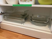 Photo showing a shelf with an assortment of clear glass and Pyrex baking pans stacked, including rectangular, square, fluted pie pans.