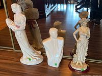 Photo showing all three sculptures: a painted cast chalk woman holding a baby, a white resin bust, and an unpainted cast chalk woman holding a baby, placed on a hardwood floor with reflections in a mirror.