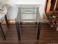 Front-facing square side table showing clear glass top and black metal base with four legs and decorative crossbars.