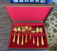 Full view of gold tone flatware set in red felt-lined wooden case showing spoons, forks, knives, and serving utensils.