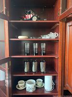 Wooden cabinet shelves with glassware, ashtrays, teacups and a candle warmer arranged on different shelves.