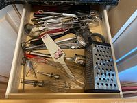 Top view of drawer with various metal and plastic kitchen utensils including whisks, ladles, peelers, spatulas, and a box grater
