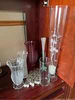 Two crystal vases, three glass vases including one tall plastic vase, and six lucite candles arranged on wood shelf