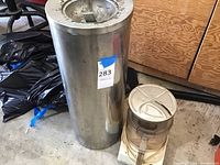 Photo of a tall cylindrical stainless steel ashtray alongside a vintage Cuisinart food processor on a floor with plywood wall background.