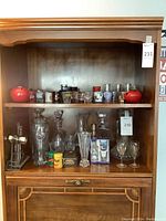 Wide photo of shelf with assorted glassware, including decanters, shot glasses, small containers, and a manual corkscrew opener.