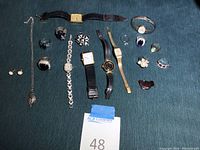 Overview of all jewelry and watches placed on a dark fabric surface in a group showing various items including rings, watches, brooches, and earrings.
